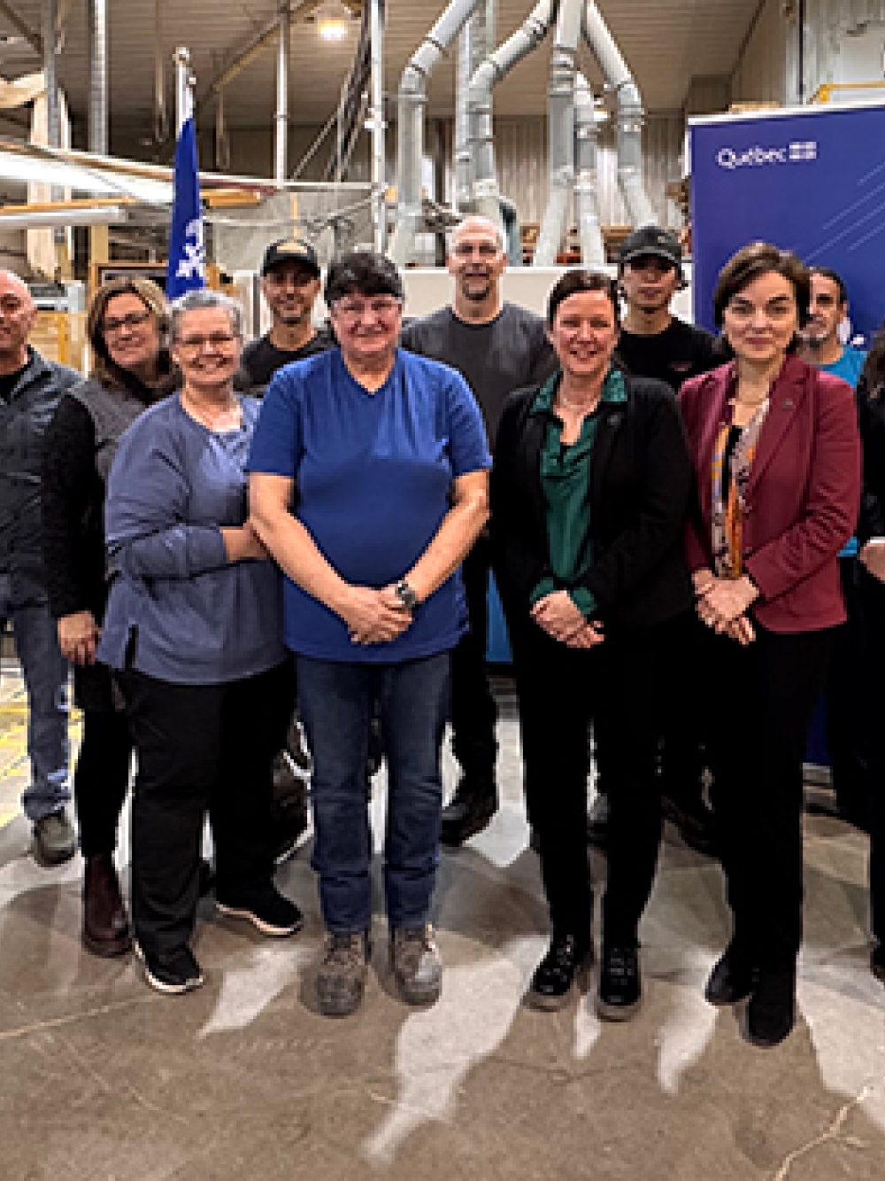 Groupe de personnes debout dans une usine, devant des équipements industriels et une bannière du gouvernement du Québec.