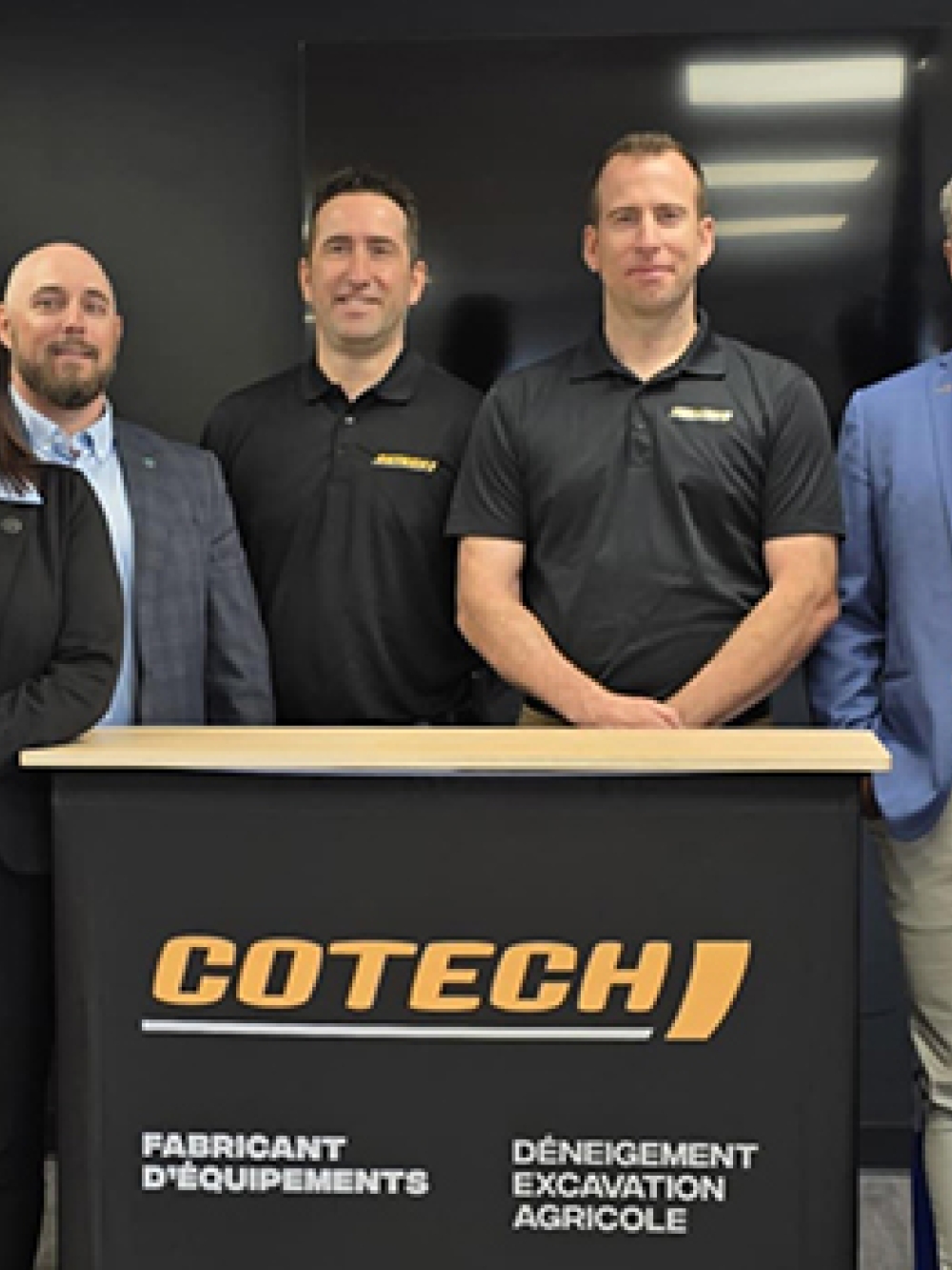 Groupe de personnes debout derrière un lutrin portant le logo de Cotech, accompagné de mentions sur la fabrication d’équipements de déneigement, d’excavation et agricoles, dans une salle de présentation.