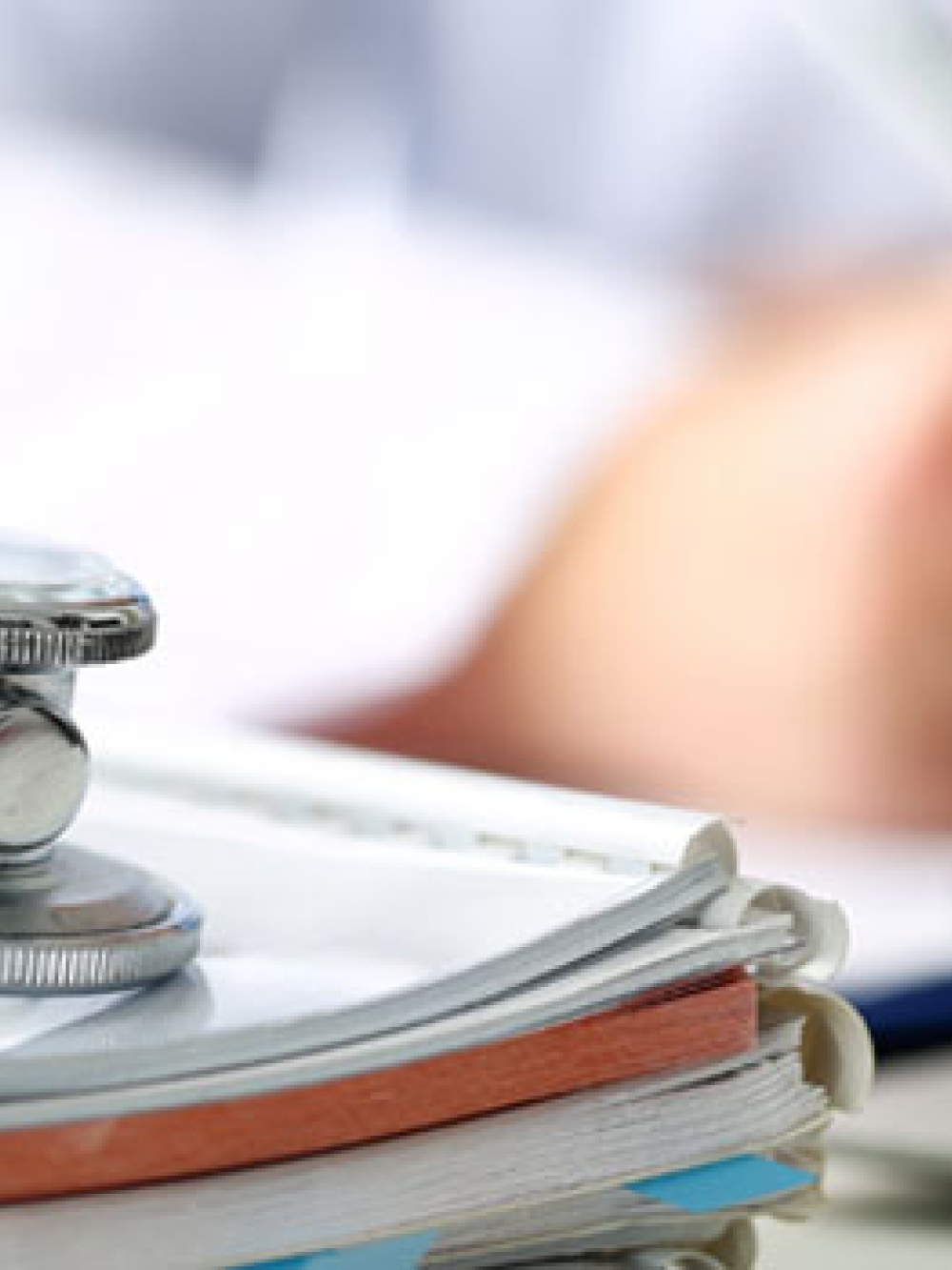 Stéthoscope posé sur une pile de documents médicaux, avec une personne en arrière‑plan écrivant à un bureau.