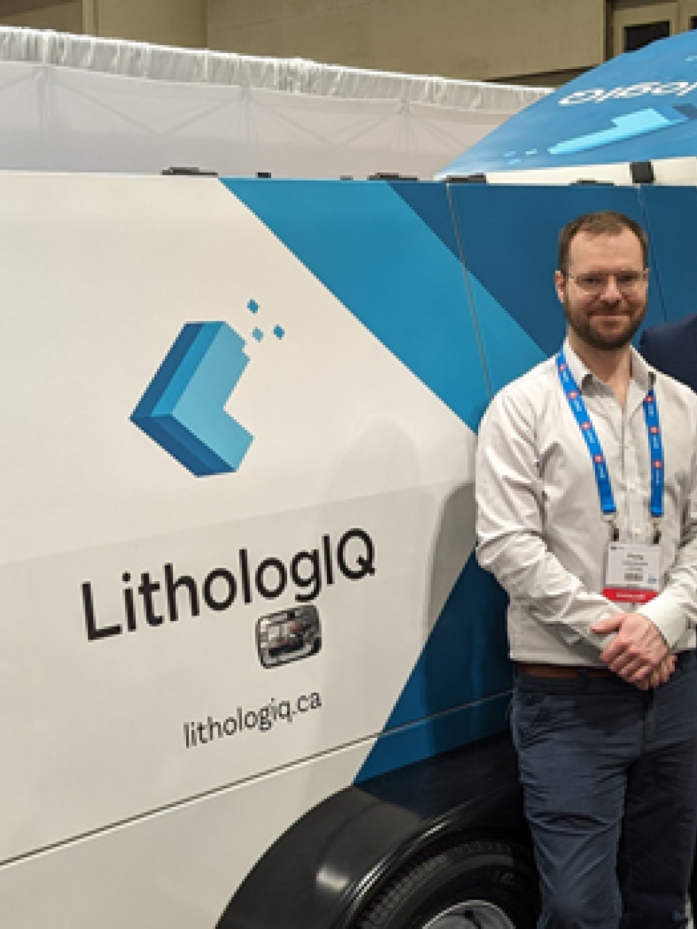 Unité mobile blanche avec le logo LithologIQ et l’inscription « Mobile Core Scanner Lab », exposée dans un salon professionnel, avec trois personnes debout à côté et des kiosques en arrière-plan.