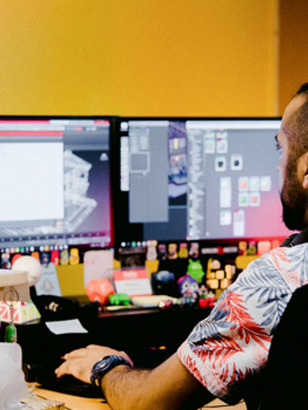 ersonne assise à un bureau dans un studio de jeux vidéo, travaillant sur deux écrans affichant des interfaces de création, avec des figurines et objets colorés autour et une chaise portant le logo Ludia.