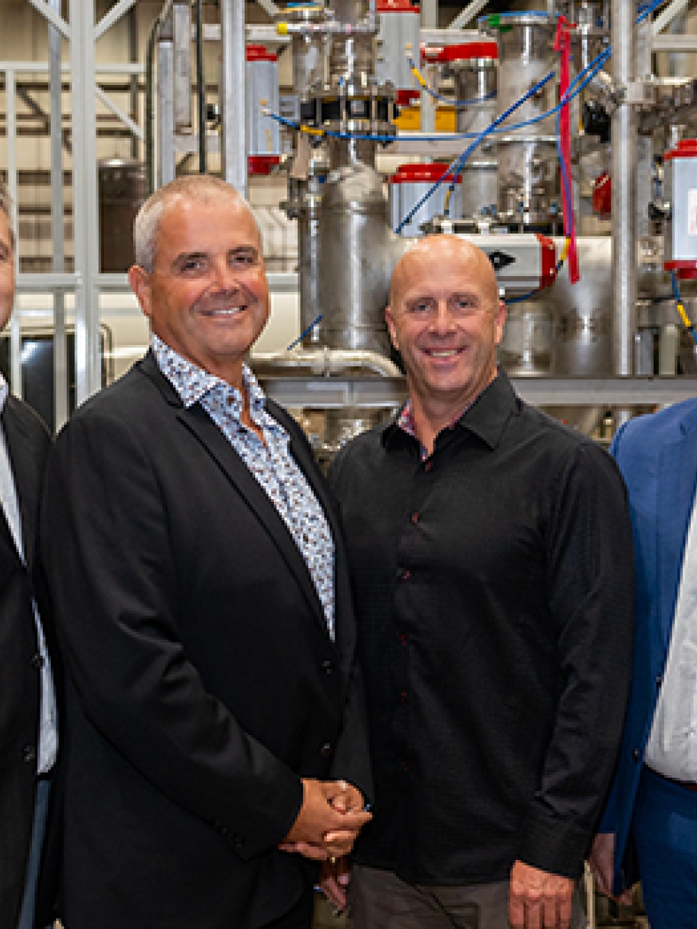 Groupe de personnes debout dans une usine, devant un ensemble de tuyaux, de valves et d’équipements industriels en acier inoxydable.