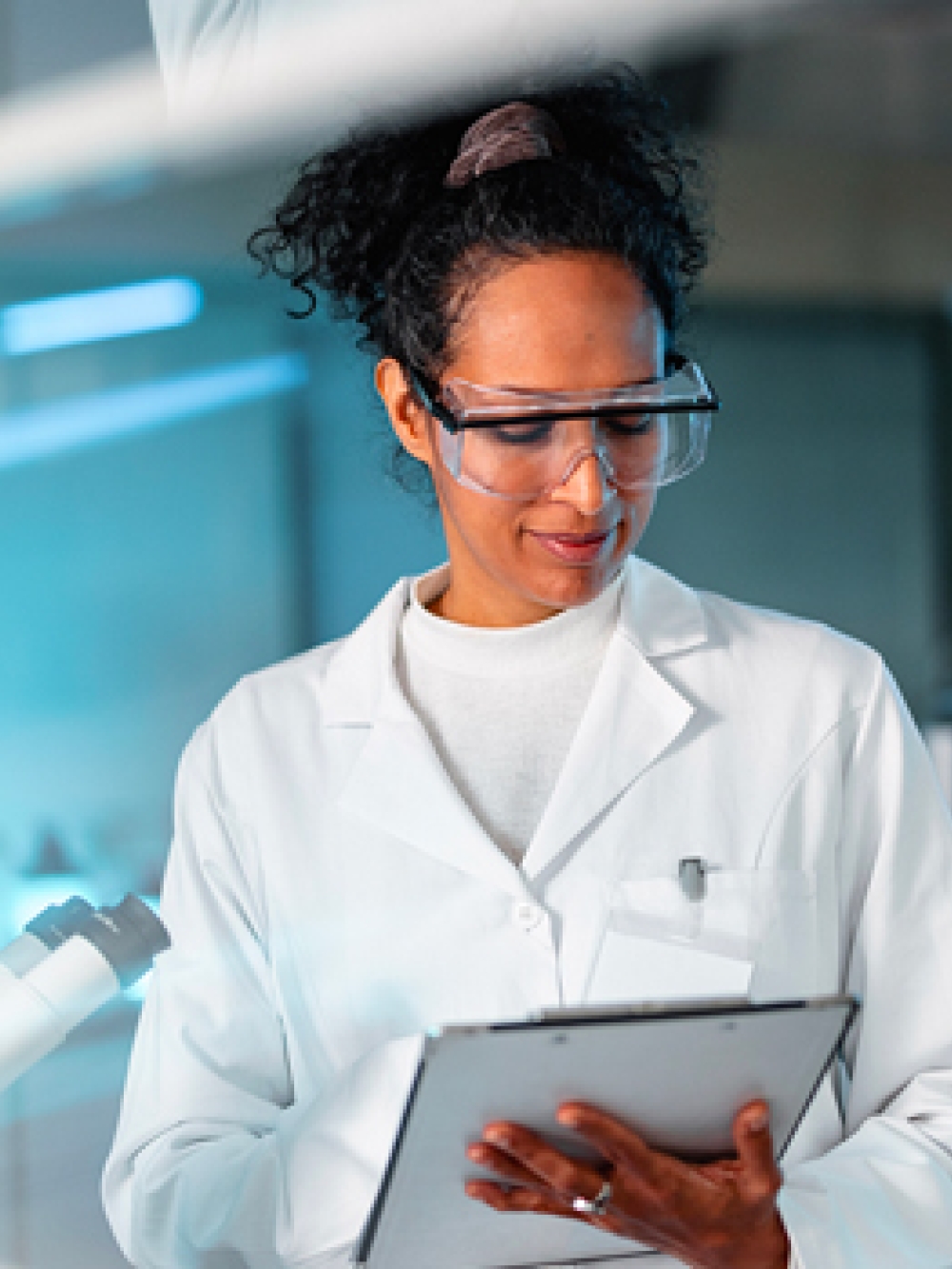 Personne en sarrau de laboratoire tenant une tablette électronique dans un environnement scientifique moderne, entourée d’équipements et de verrerie.
