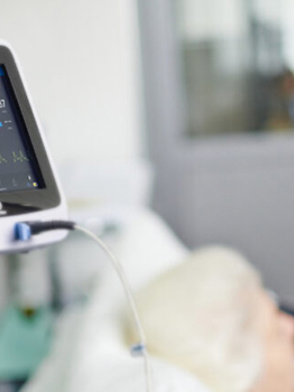 Appareil médical affichant des données de surveillance posé près d’un patient alité, avec un professionnel de la santé en arrière‑plan.