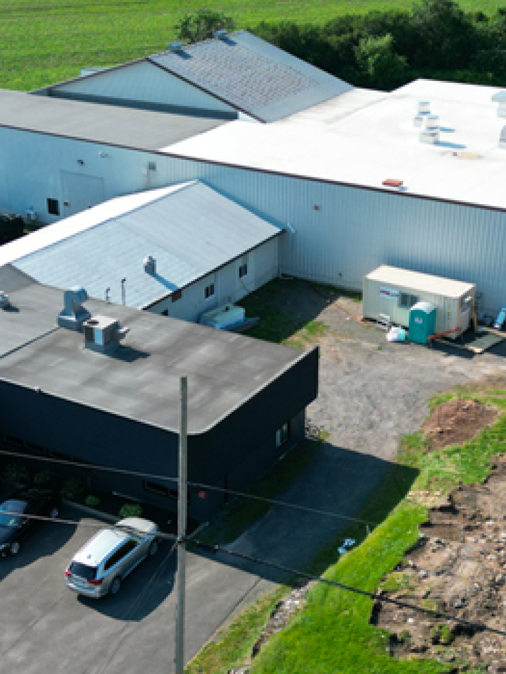 Vue aérienne d’une usine avec plusieurs bâtiments, stationnements et véhicules, montrant un agrandissement en cours avec une zone de construction et un chariot élévateur orange.