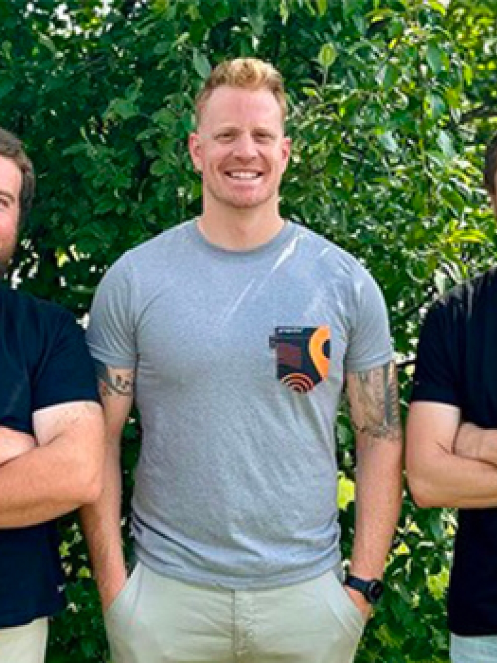 Trois personnes debout à l’extérieur devant un feuillage vert, bras croisés.