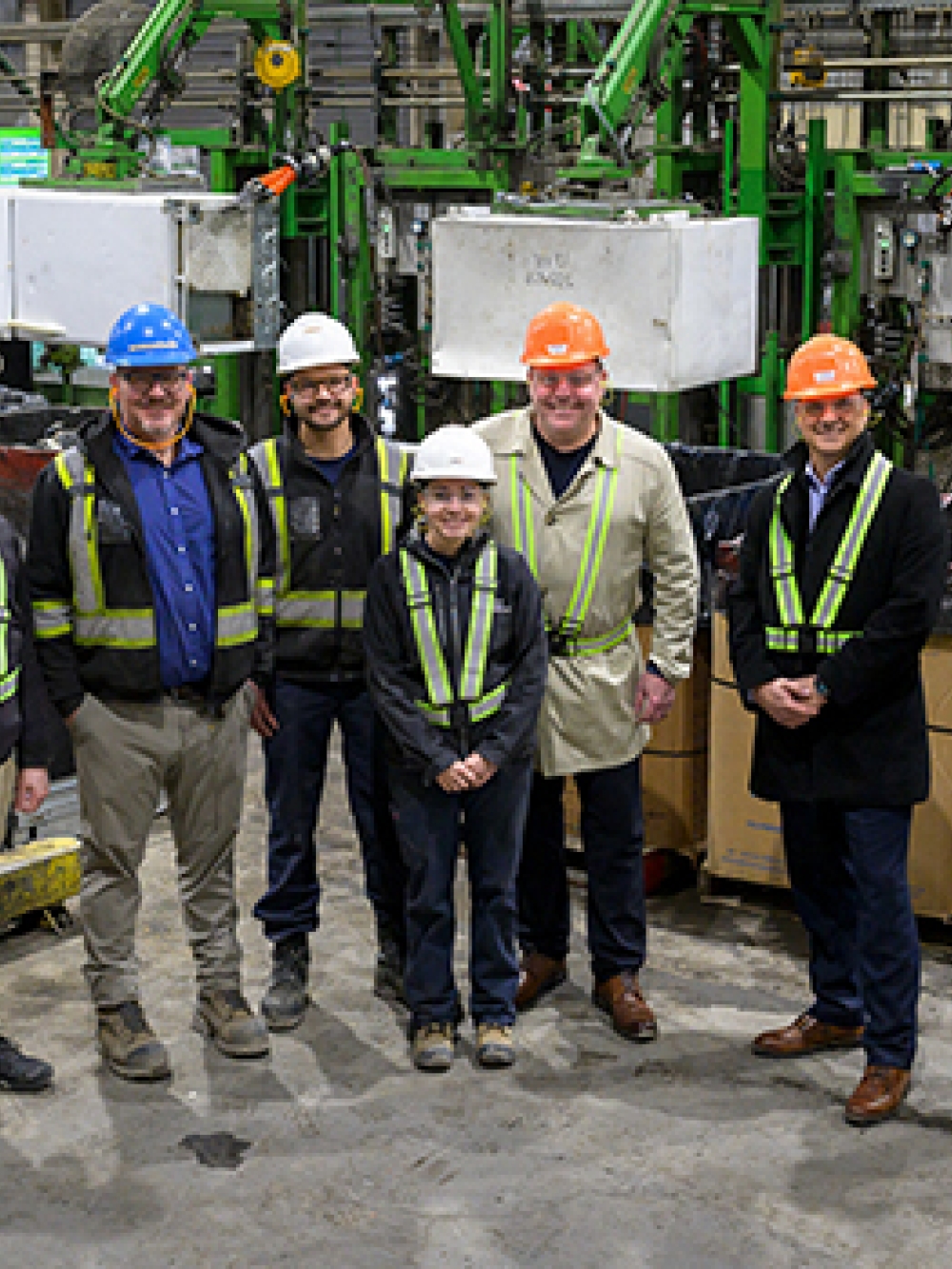 Groupe de personnes portant des casques et des équipements de sécurité dans une usine, posant devant des machines industrielles.