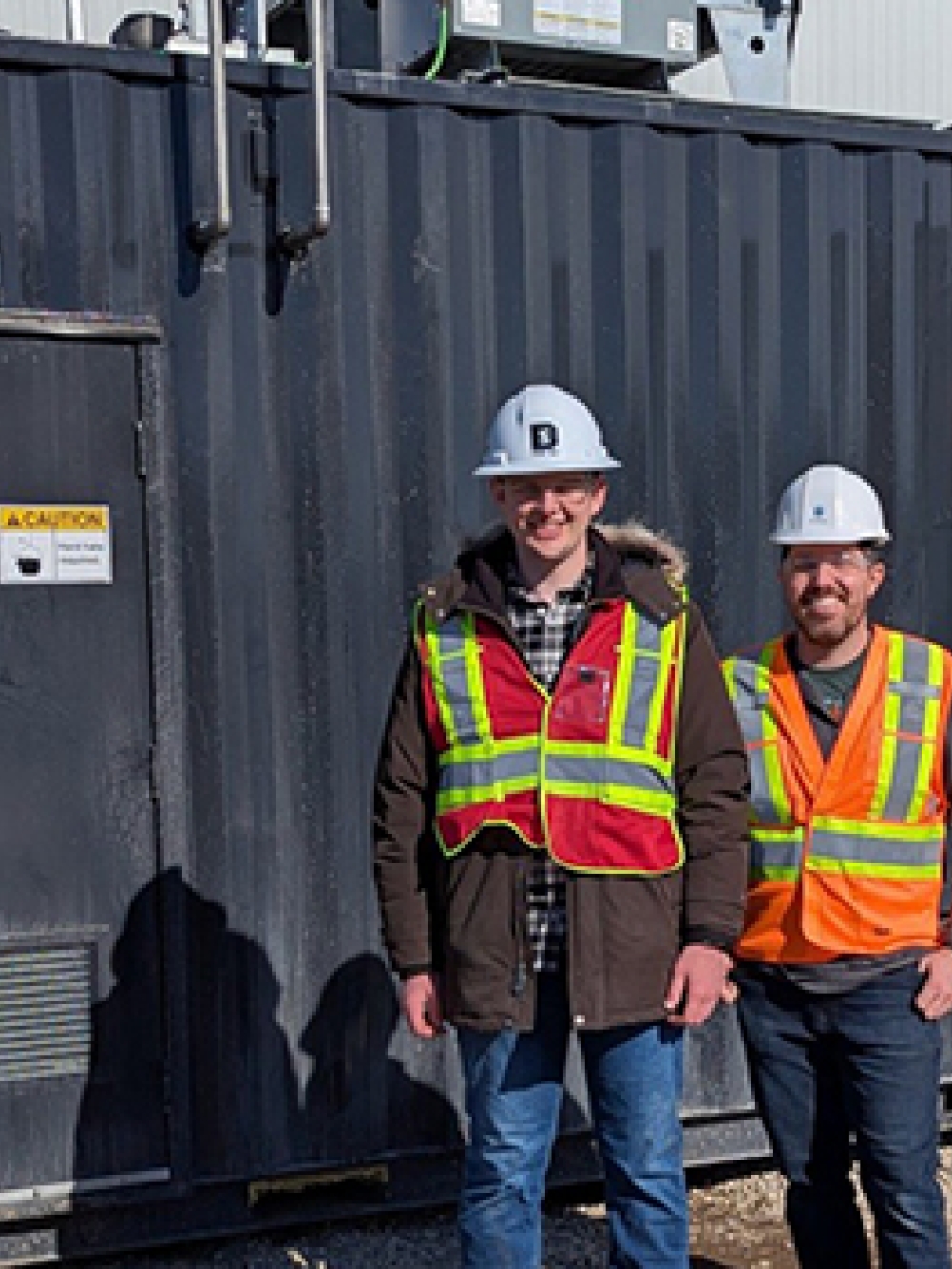 Trois personnes portant des casques de sécurité et des vestes haute visibilité debout devant une unité industrielle noire portant l’inscription « SKYRENU 50 » sur un site extérieur.