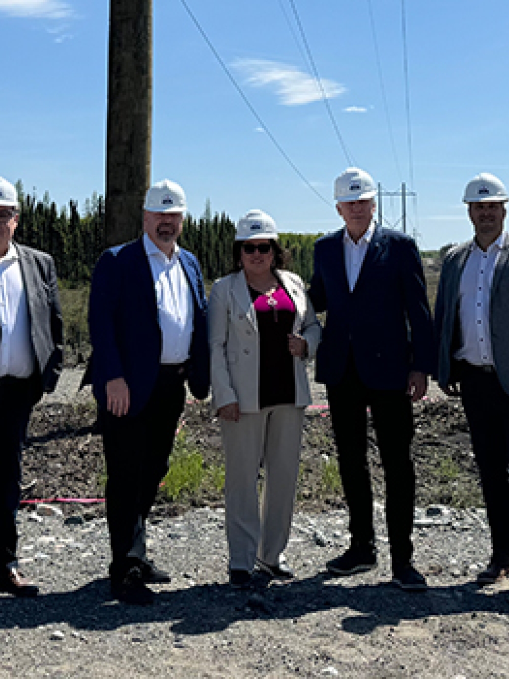 Groupe de personnes en tenue professionnelle et casques de sécurité posant à l’extérieur sur un site de construction, avec des poteaux électriques et des lignes de transmission en arrière-plan.