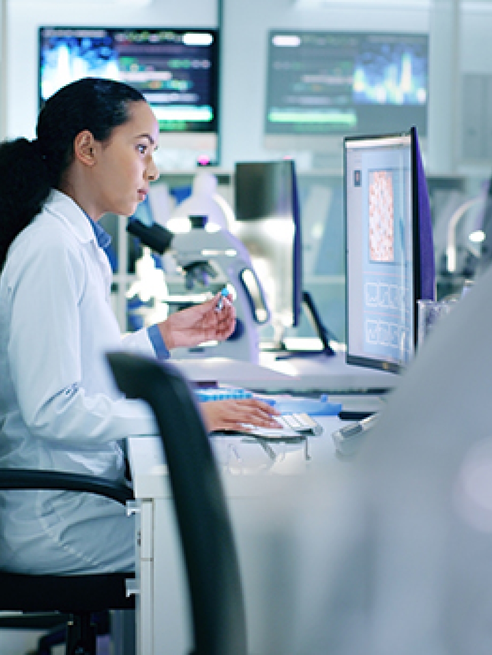 Des personnes en blouse de laboratoire travaillent sur des ordinateurs et des microscopes dans un environnement de recherche moderne, entouré d’écrans d’analyse et d’équipements scientifiques. 
