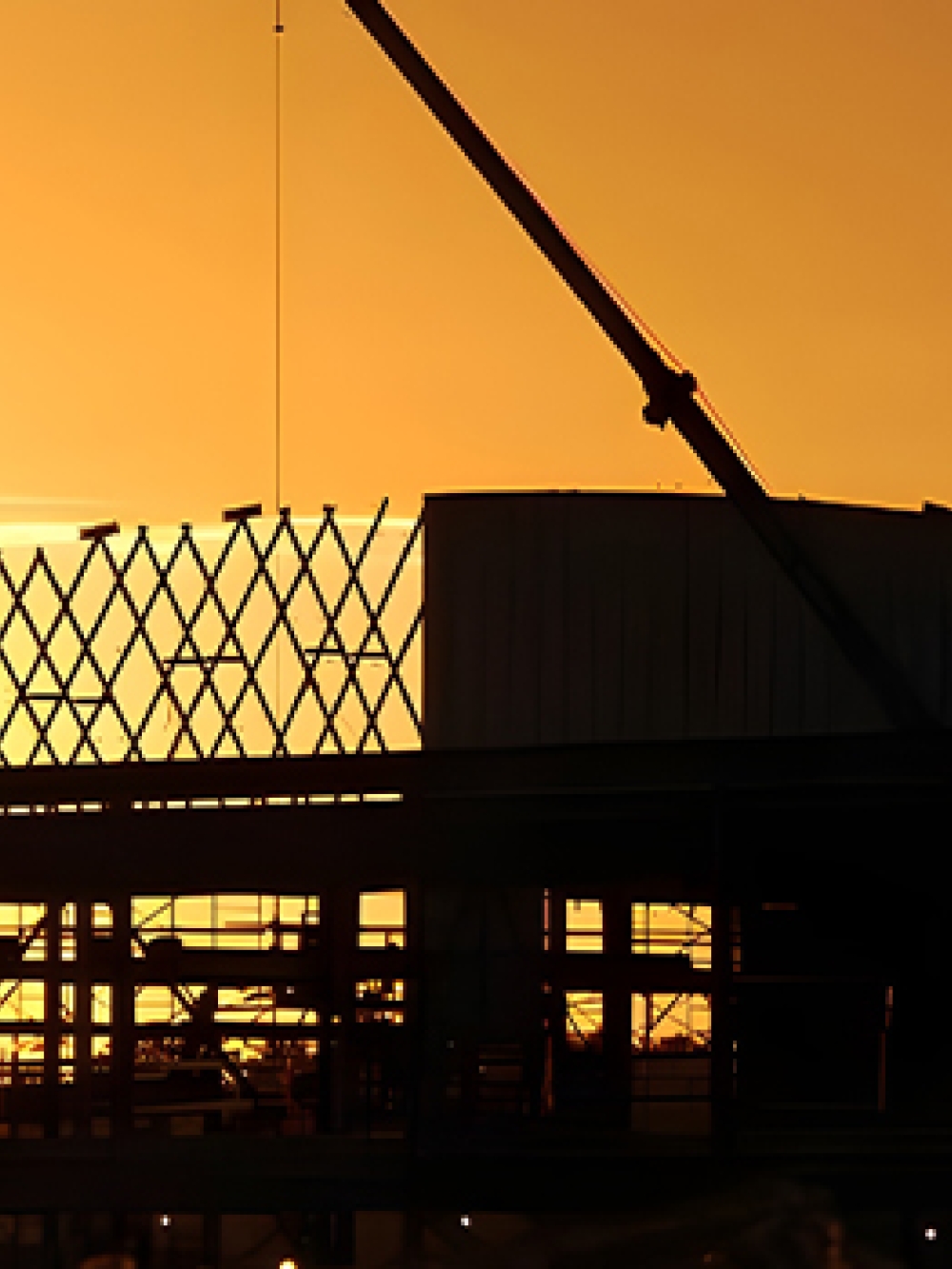 Structure métallique d’un bâtiment en construction, silhouettée devant un ciel orange au coucher du soleil, avec une grue visible au-dessus du chantier.