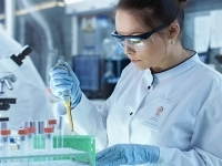 Personne en blouse blanche et gants bleus manipulant une micropipette dans un laboratoire, avec des tubes à essai dans un support vert, un microscope et divers équipements scientifiques en arrière-plan.