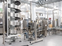 Salle de production industrielle avec des équipements automatisés pour l’embouteillage et le conditionnement, incluant des convoyeurs, des machines en acier inoxydable et des conduits aériens, dans un environnement propre et lumineux.