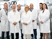 Groupe de personnes portant des sarraus blancs dans un laboratoire moderne, entouré d’équipements scientifiques, d’étagères avec produits et d’un grand réfrigérateur vitré.