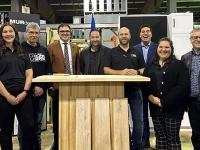 Groupe de personnes debout derrière un lutrin en bois dans un atelier industriel, avec des équipements et matériaux de scierie visibles en arrière‑plan ainsi qu’un écran de présentation sur la gauche.