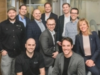 Groupe de personnes posant dans un bureau moderne, devant une paroi vitrée où l’on aperçoit une salle de réunion et des tableaux avec des notes.