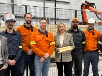 Groupe de neuf personnes posant dans une usine, dont plusieurs portent des vêtements de travail orange ou foncés, avec des équipements industriels et des robots derrière une grille de sécurité.