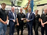 Groupe de personnes debout dans un atelier industriel, entourées de machines-outils et d’équipements de fabrication, certaines tenant des pièces mécaniques métalliques.