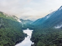 Vue aérienne d’une vallée verdoyante avec une rivière sinueuse entourée de montagnes boisées et de nuages bas.