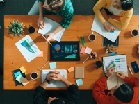 Vue aérienne d’un groupe de personnes autour d’une table de travail, échangeant sur des graphiques, documents et un écran affichant des données.