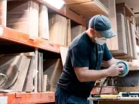 Un travailleur dans un atelier industriel, en train de poncer une pièce de bois sur un établi, entouré d’étagères contenant des panneaux et matériaux empilés.