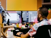 ersonne assise à un bureau dans un studio de jeux vidéo, travaillant sur deux écrans affichant des interfaces de création, avec des figurines et objets colorés autour et une chaise portant le logo Ludia.