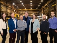 Groupe de personnes debout dans un grand atelier industriel, posant entre des équipements, des structures métalliques.