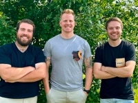 Trois personnes debout à l’extérieur devant un feuillage vert, bras croisés.