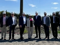Groupe de personnes en tenue professionnelle et casques de sécurité posant à l’extérieur sur un site de construction, avec des poteaux électriques et des lignes de transmission en arrière-plan.